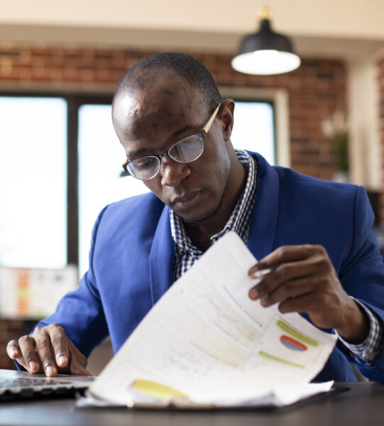 Closeup of black man seated at office desk, deeply focused on analyzing financial documents in brick wall room. African american male manager examining graphs on clipboard with laptop nearby.